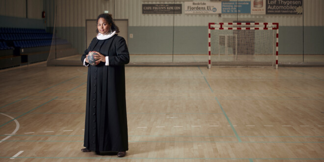 Kvindelig præst iført præstekjole i tom håndboldhal med håndbold i hænderne