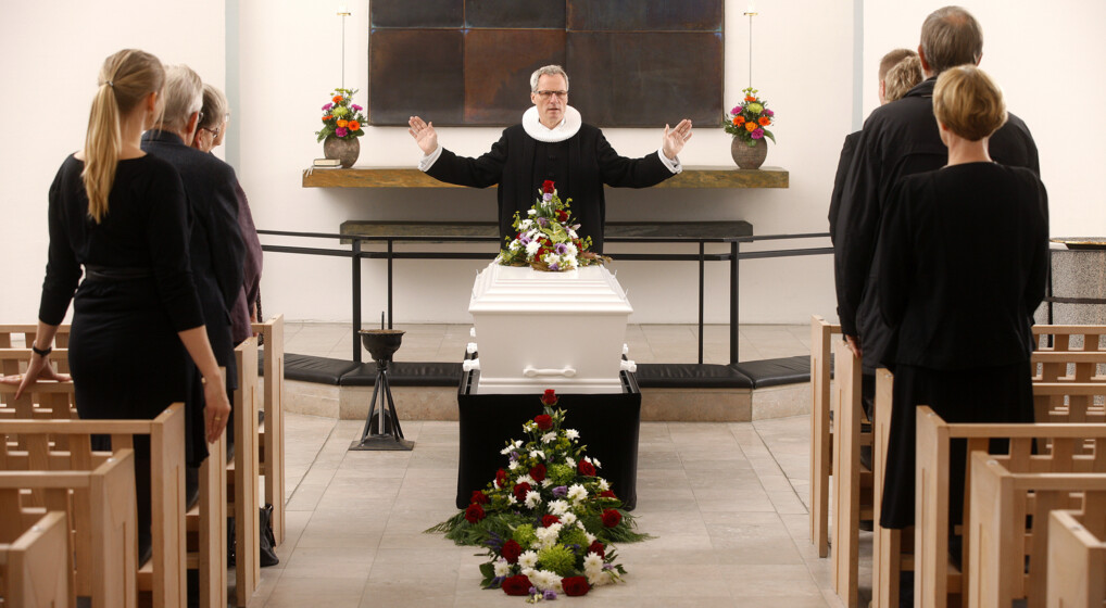 En præst lyser velsignelsen foran en hvid, blomsterpyntet kiste i en kirke.
