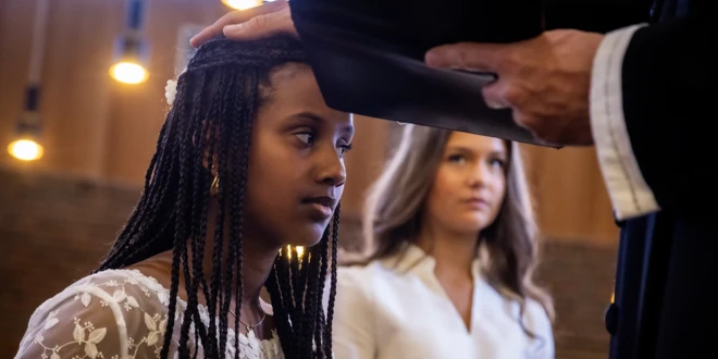 En pige i konfirmationskjole knæler, mens præsten lægger hånden på hendes hoved og konfirmerer hende.