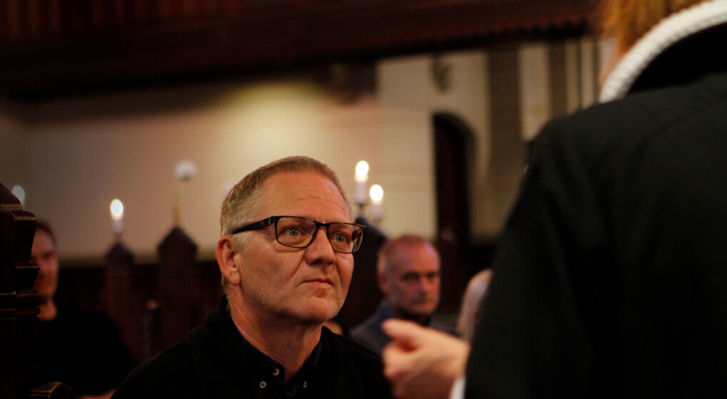 Voksen mand og præst ved døbefonten til drop in-dåb med mennesker i baggrunden, der venter på at blive døbt.