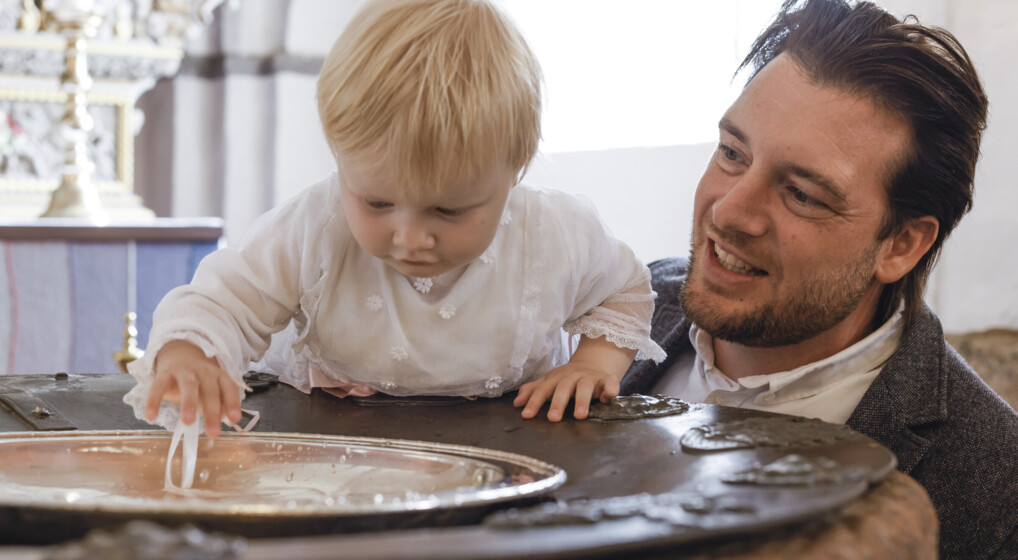 Dåbsbarn leger med vandet i døbefonten, mens far ser til