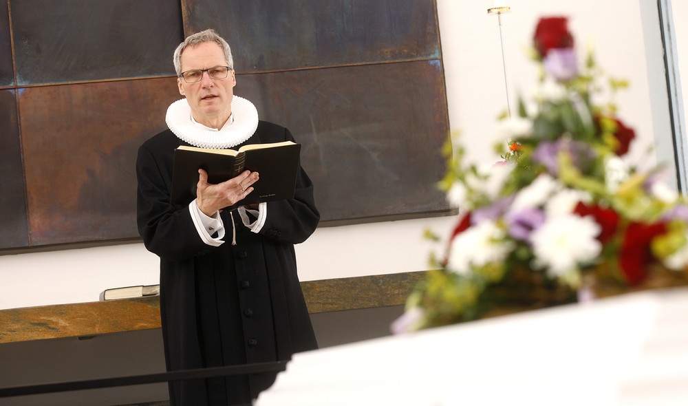 Præst står foran alteret, bag kiste og læser højt fra ritualbogen