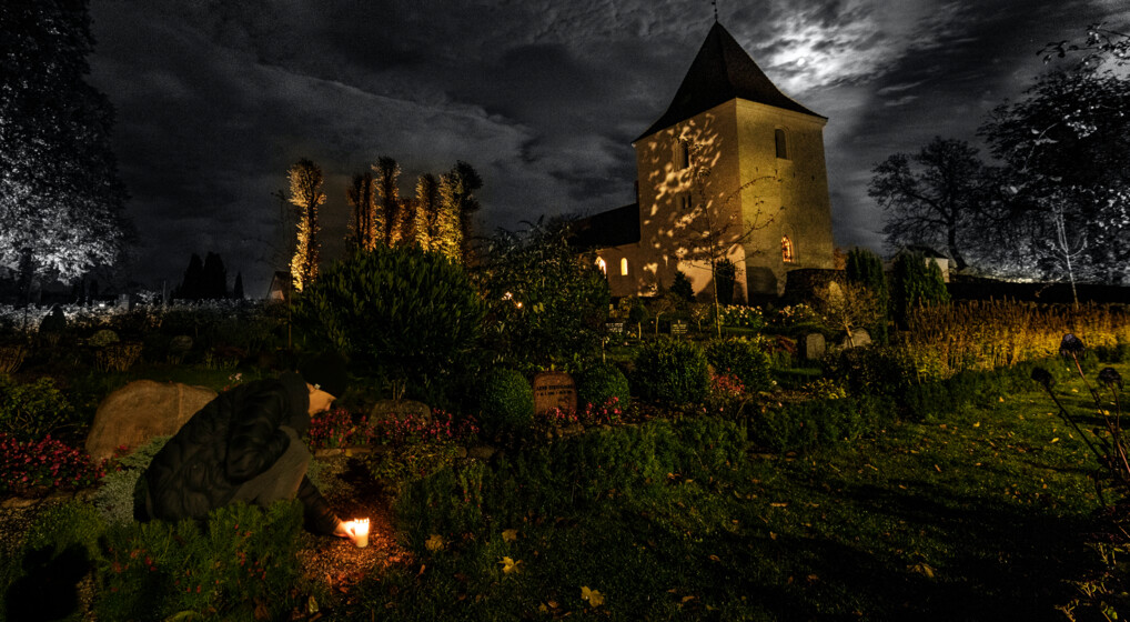 Billede af kirkegård i tusmørke, hvor en person tænder et gravlys