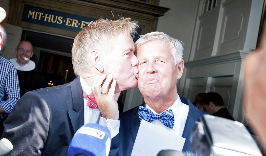 En mand kysser sin nylige ægtemand på kinden inde i kirken, mens præsten i baggrunden smiler