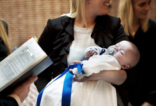 Smilende mor holder baby i dååbskjole med blå slløjfe og sut