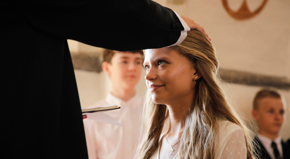 En pige i hvid konfirmationskjole bliver konfirmeret ved at præsten lægger sin hånd på hendes hoved.