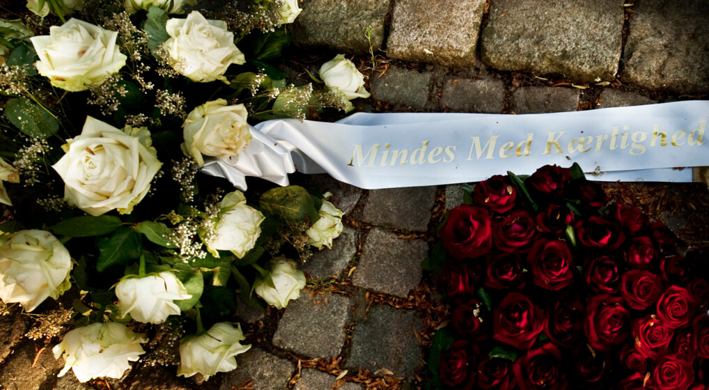 To bårebuketter og et hvidt bånd med på skriften 'Mindes med kærlighed' ligger på jorden.
