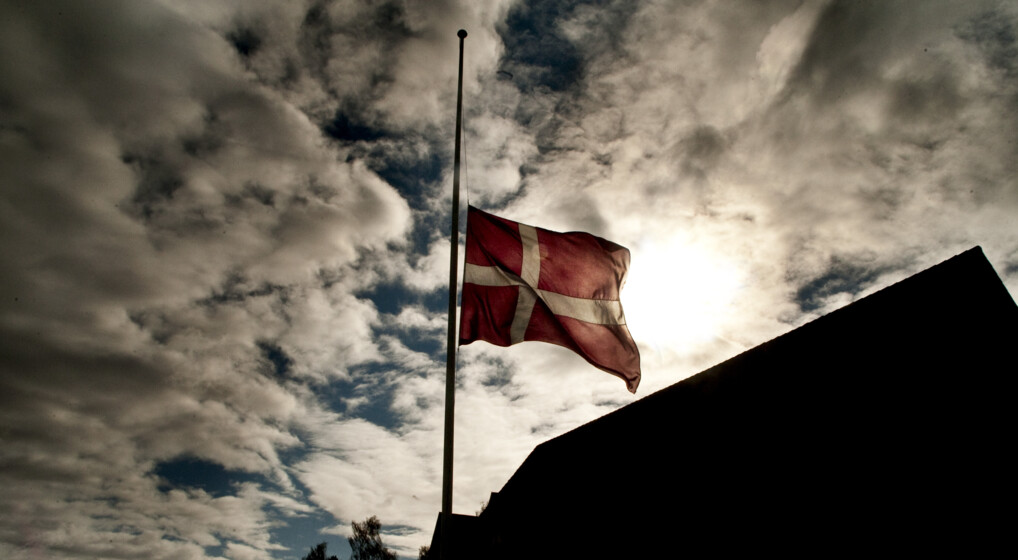 Flag på halvt uden for en kirke. Bag flaget ses solen gemt bag skyerne.